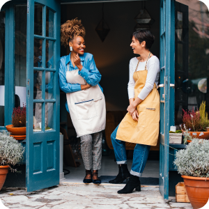 Two female small business owners standing in the doorway of their shop talking to each other happily.