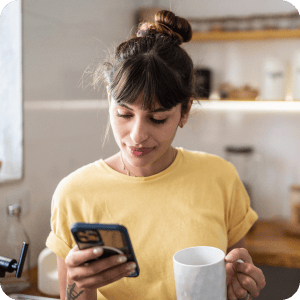 Woman in a yellow shirt holding a cup of coffee and looking at her cell phone.