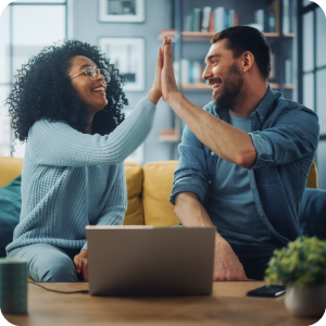 Two friends sitting on a couch in front of a computer giving each other a high five.